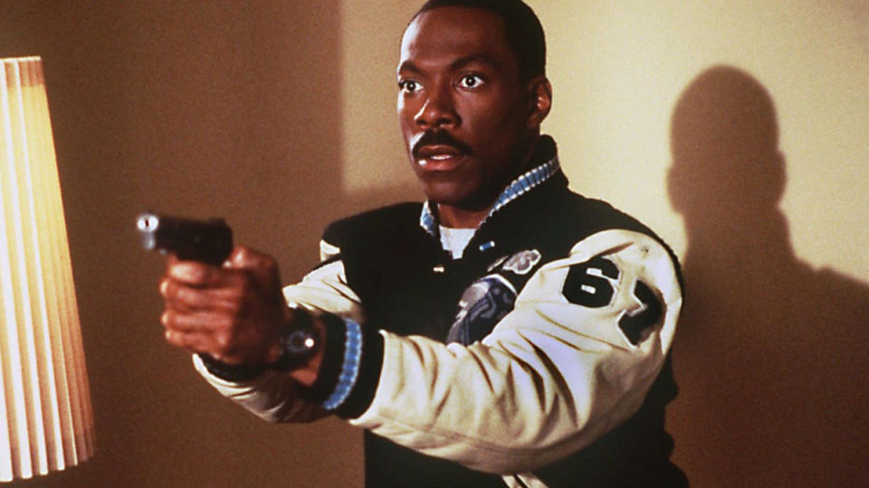 Eddie Murphy as detective Axel Foley holding a gun and wearing a black jacket with white sleeves.