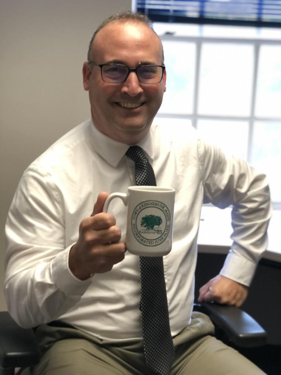Stephen Houdlette is the new director of economic vitality for the town of Kennebunk. He is seen here, lifting a mug sporting the town's trademark Lafayette Elm, in his office at the town hall on June 29, 2023.