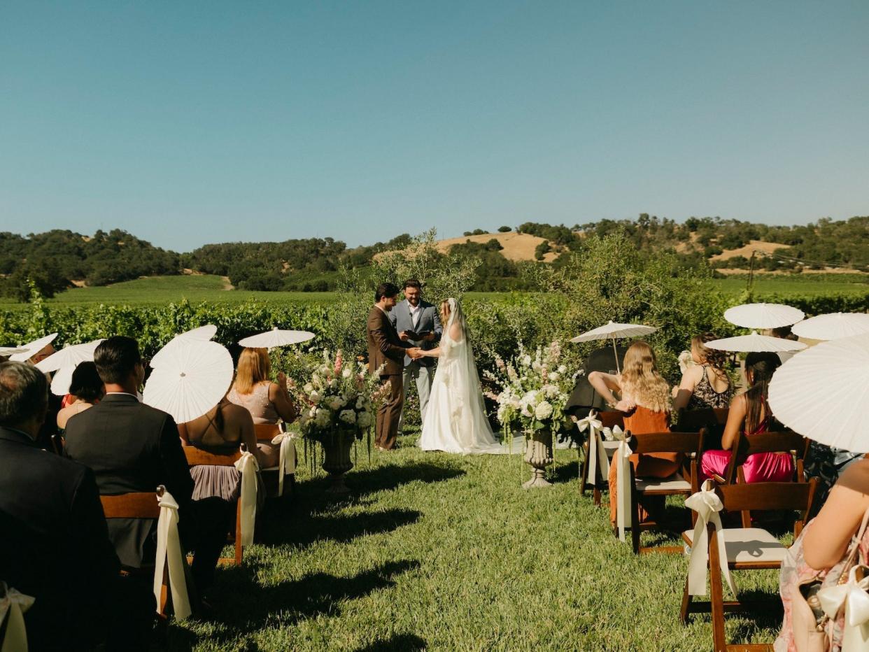 A bride and groom hold hands during their wedding ceremony in a vineyard, with guests on either side of the aisle holding parasols.