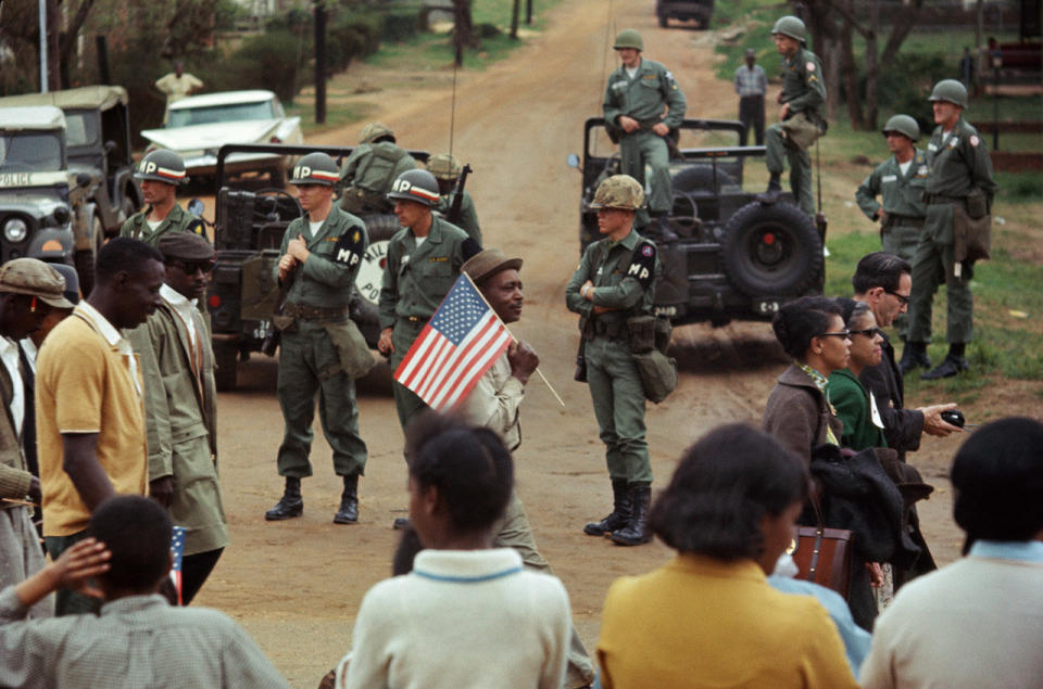 Man with American flag and marchers walking past federal troops guarding crossroads, 1965.