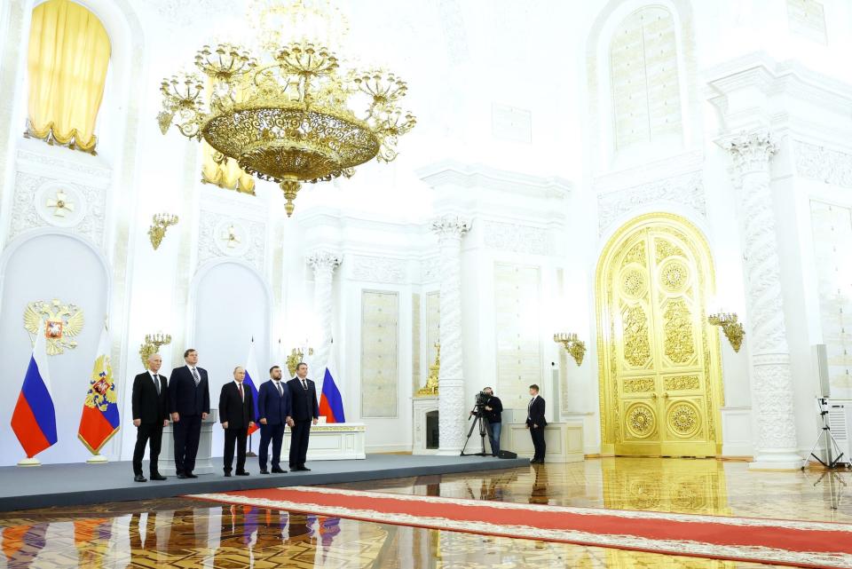 Russian President Vladimir Putin stands in a white and gold gilded room with officials on either side of him