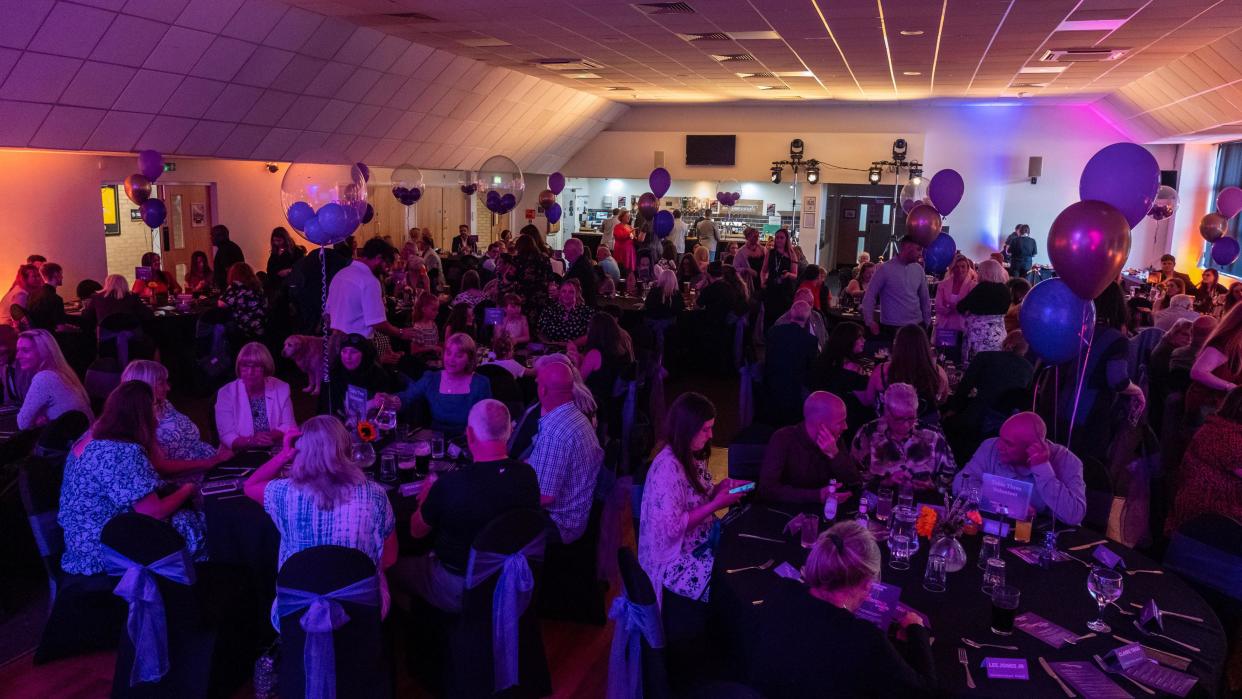 More than 150 people sat inside a function room on round tables. The room is darkly lit and balloons are on each table