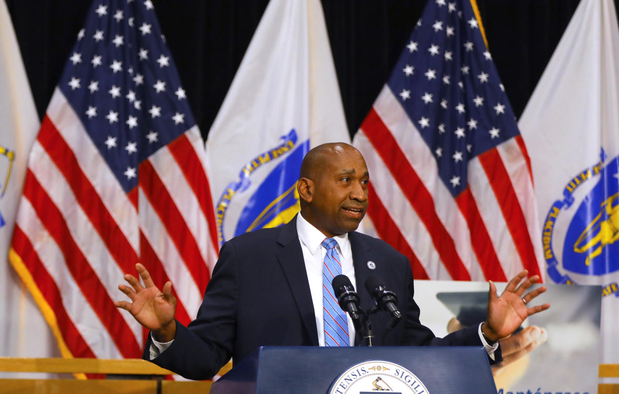 Rep. Russell E. Holmes stands at a podium with hands raised, palms out, in front of flags.