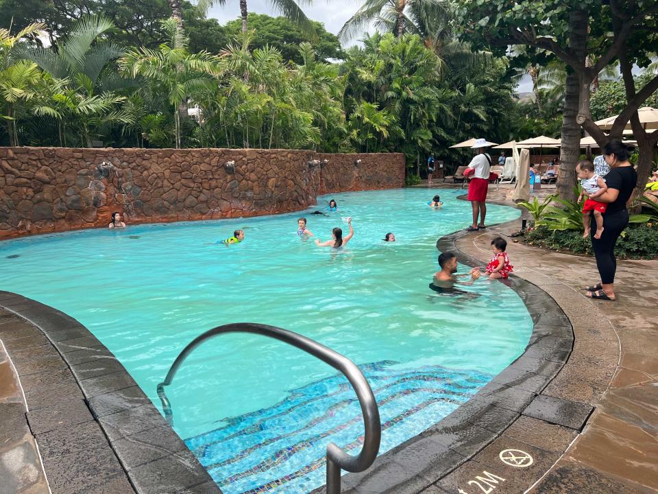 A long pool with a wall featuring waterfalls, surrounded by lush trees, with a lifeguard watching over.