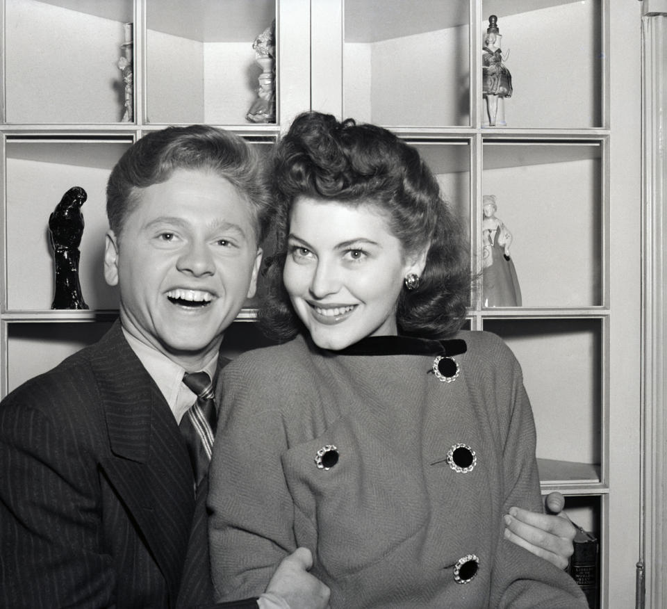 Mickey Rooney and Ava Gardner smile as they pose in front of a shelf of figurines. Mickey wears a pinstripe suit and tie; Ava wears a buttoned coat