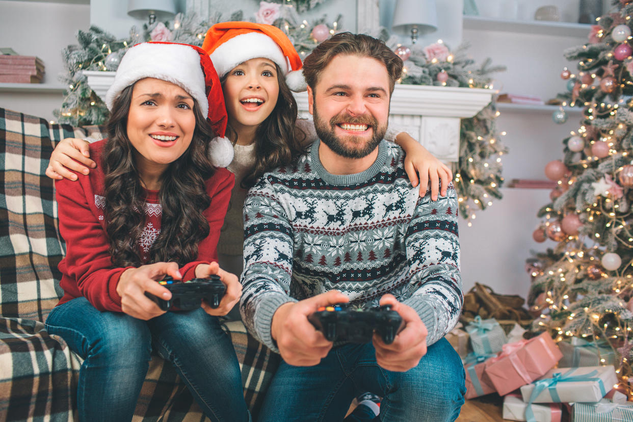 Playful and happy family sitting on sofa and playing games using joystick. Child is between parents. Young man and woman playing games. They look festive