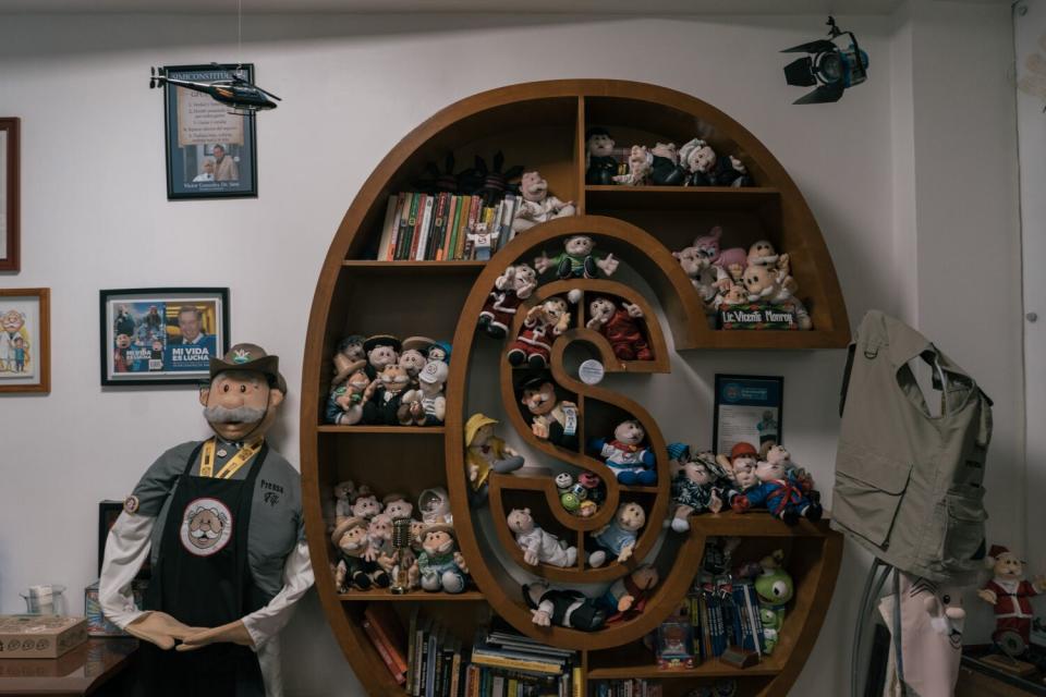 Plush dolls sit on a bookcase in the shape of the letters C and S, next to a larger version of the mustachioed character