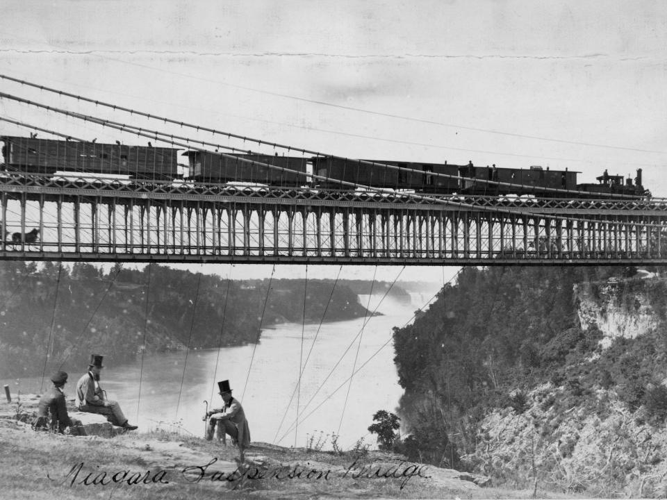 Men sit on Niagara cliffs as a steam train and a horse and carriage cross on the two-tier suspension bridge