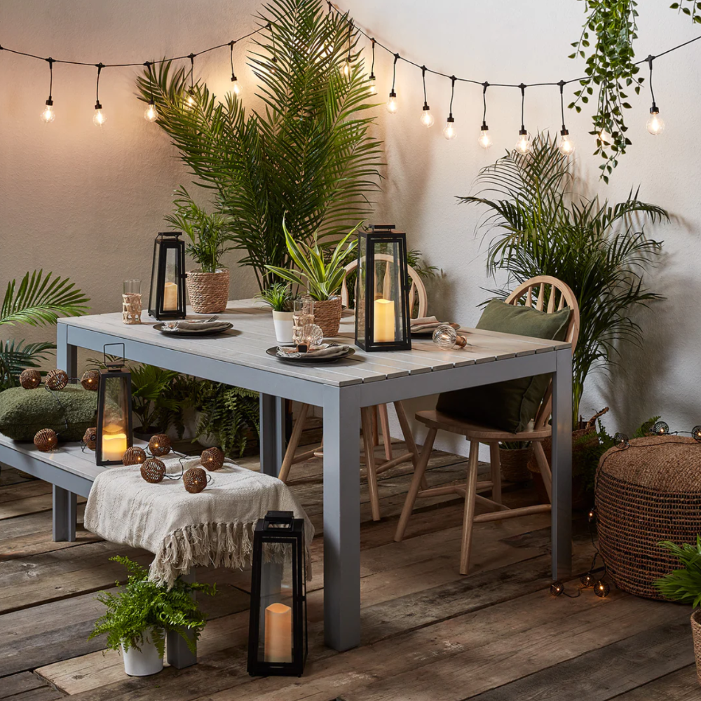  Photo of grey table surrounded by green plants, cancles, and hanging lights. 