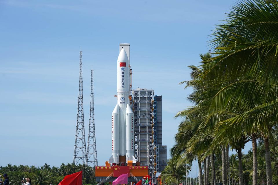 large white rocket with red markings rolls down tree lined avenue with large metal assembly building in the background