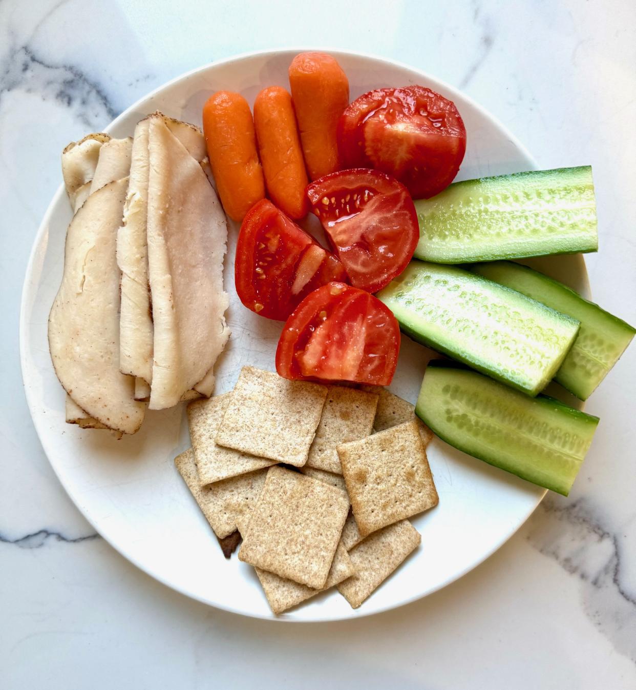 A plate of sliced turkey, baby carrots, sliced tomatoes, sliced cucumber and crackers.