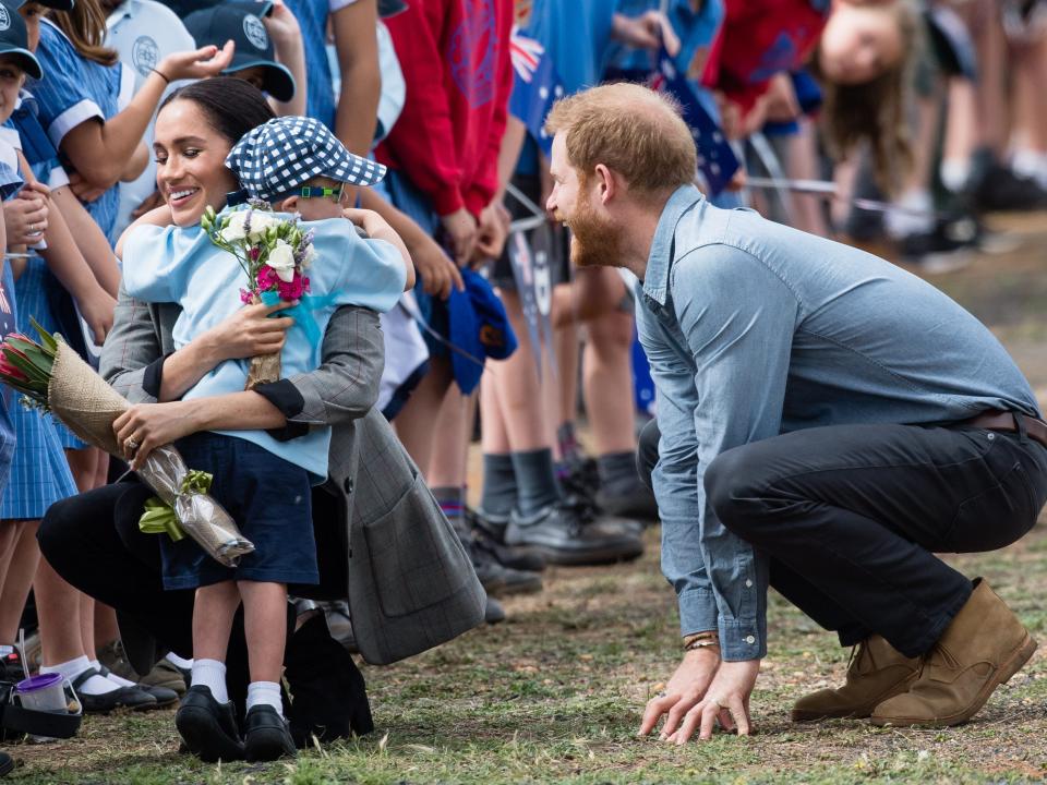 Markle squatting and smiling while hugging a young boy. Harry also squats in a frog position with his hands on the ground and smiling at the pair hugging.