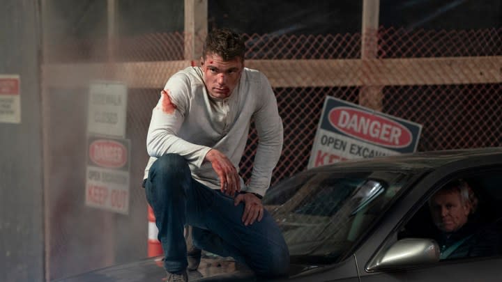 Peter kneeling on top of a car, bloody and battered in a scene from The Night Agent.