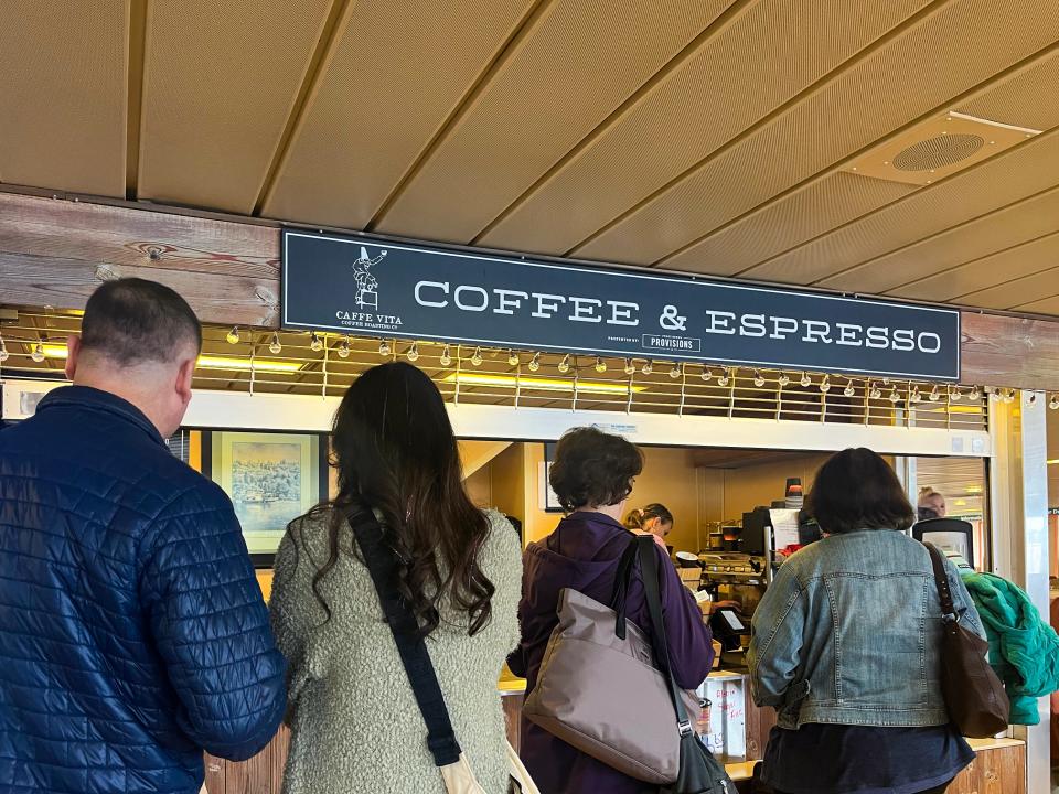 Four people in line at a ferry coffee shop with a black sign reading "Coffee & Espresso"