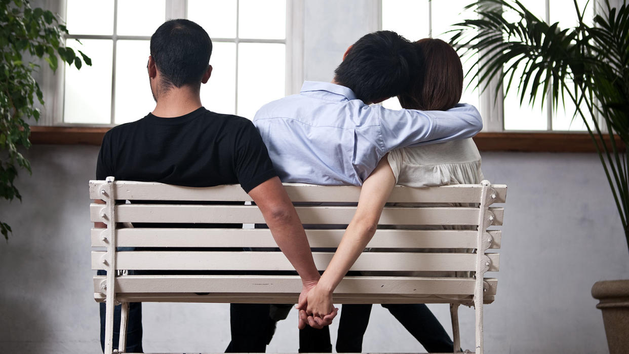 Photo illustrating infidelity, with a woman holding hands with one man while being hugged by another.