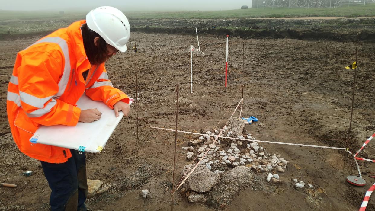  an archaeologist in an orange jacket bent over a tablet and sketching. beside them on the ground is a set of stones in an approximatel circular shape. the area is marked off and protected by small poles and strings  