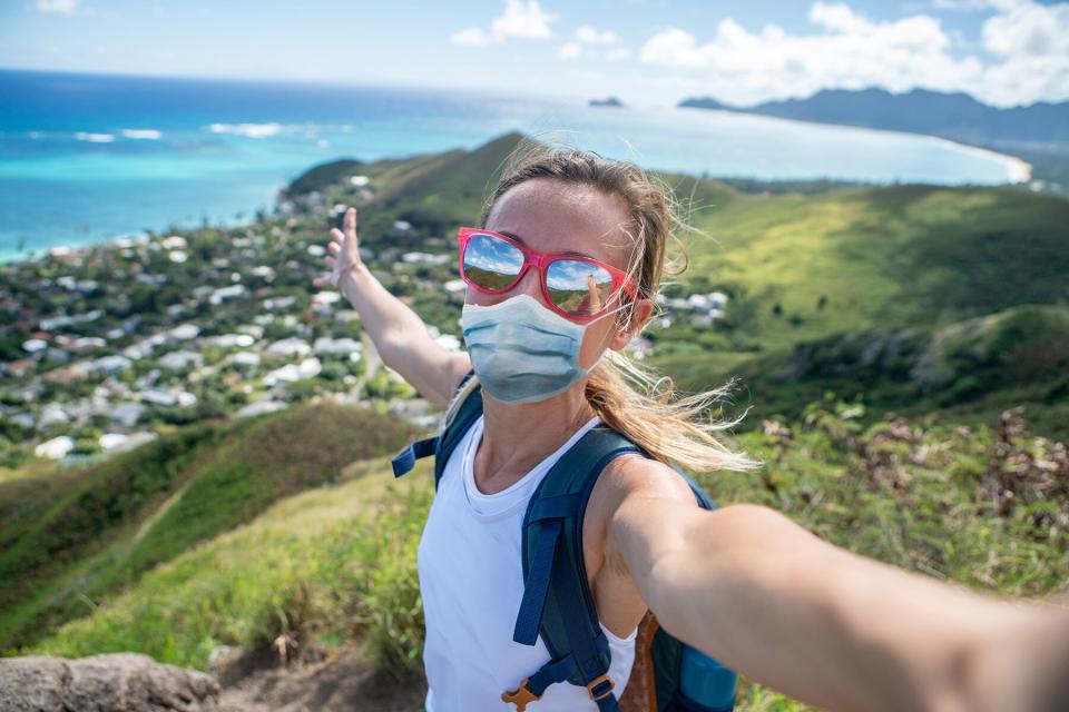 Woman in a facemask takes selfie on cliff overlooking coastline of Hawaii