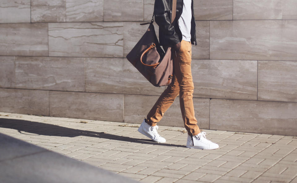 Fashionable man in black rock leather jacket with bag walking over grey textured at evening city background