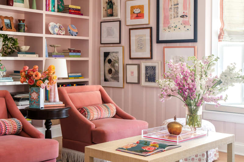 Two fringed pink chairs in front of light coffee table with flowers in pink library with gallery wall and built-in shelves.