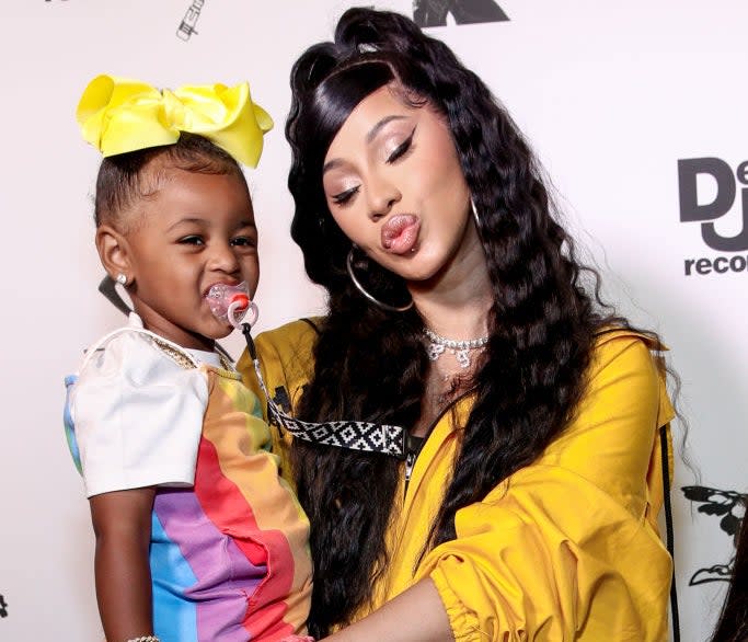 Cardi B in a draped outfit holding her daughter, who's in a colorful dress with a yellow bow in her hair