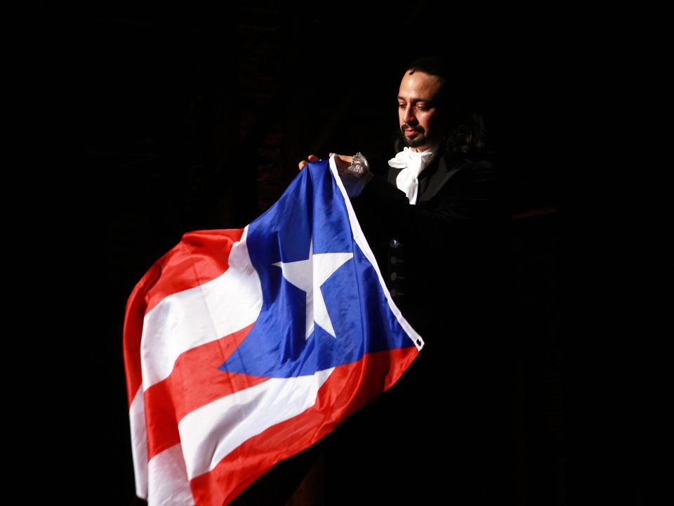 Lin-Manuel Miranda dressed in character as Alexander Hamilton waving a Puerto Rican flag on a dark stage.