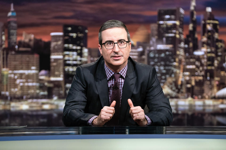 This March 3, 2019 photo released by HBO shows John Oliver, host of "Last Week Tonight with John Oliver." Six people at CBS News have tested positive for the coronavirus. Oliver tapes in a studio in the New York complex, and there were also people in the HBO show's office that tested positive. So Oliver on Sunday night taped a show without an audience from a studio where he sat in front of a white backdrop. (Lloyd Bishop/HBO via AP)