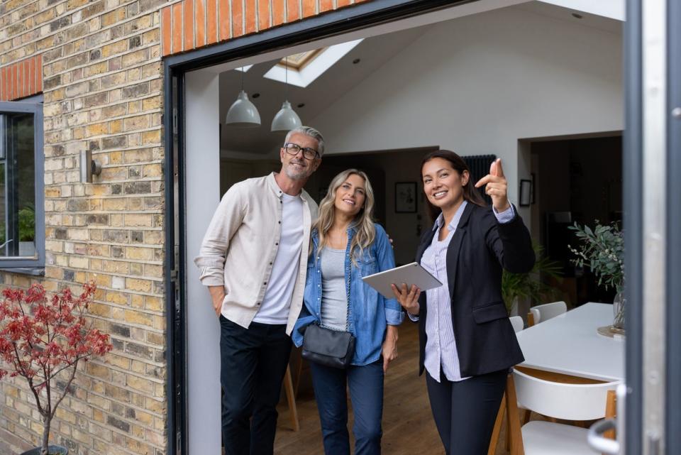 Three people smile in the entrance of a house while one woman points toward the distance. 