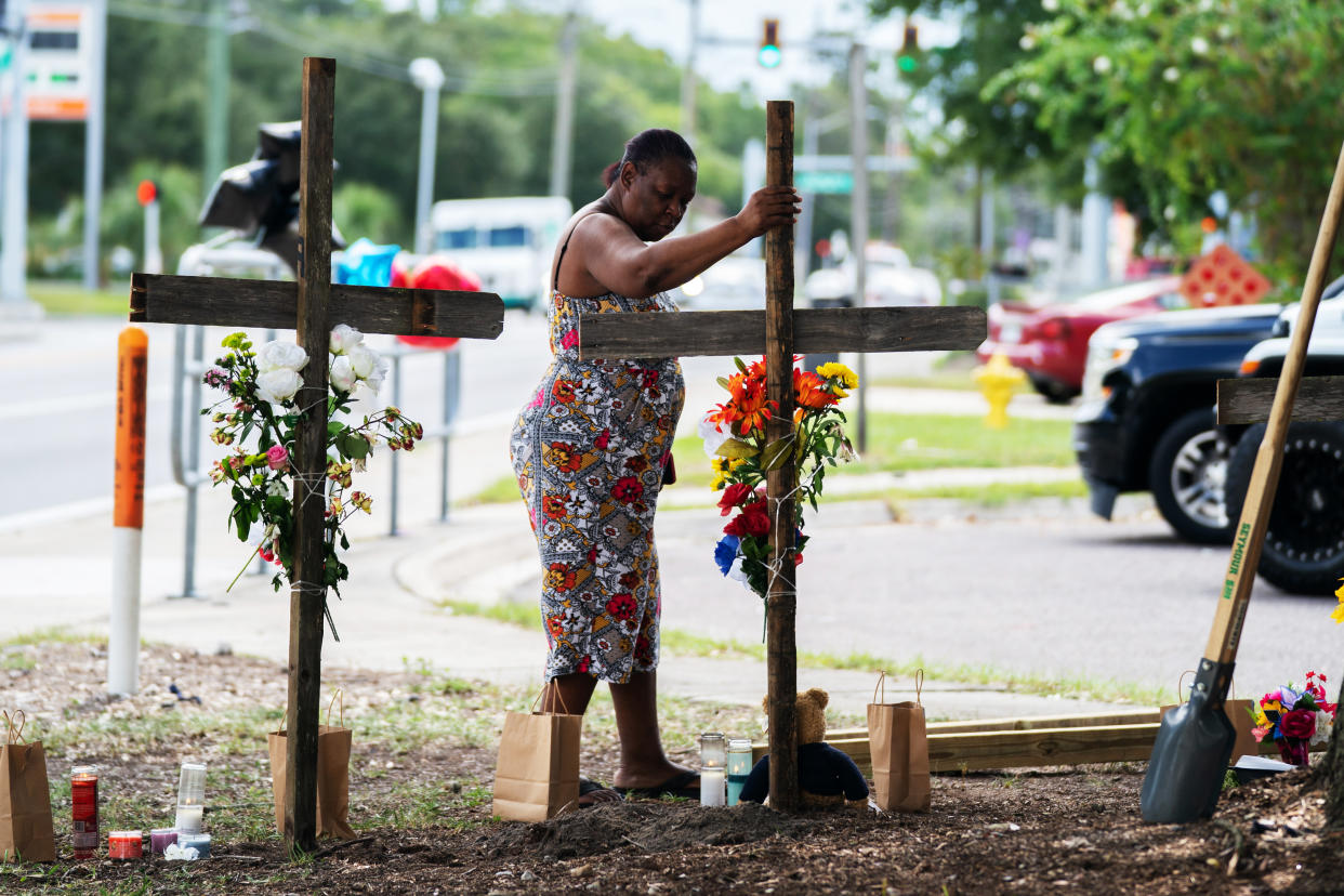 A person stands by a sidewalk near a road next to two wooden crosses decorated with flowers alongside candles and a post hole digger.
