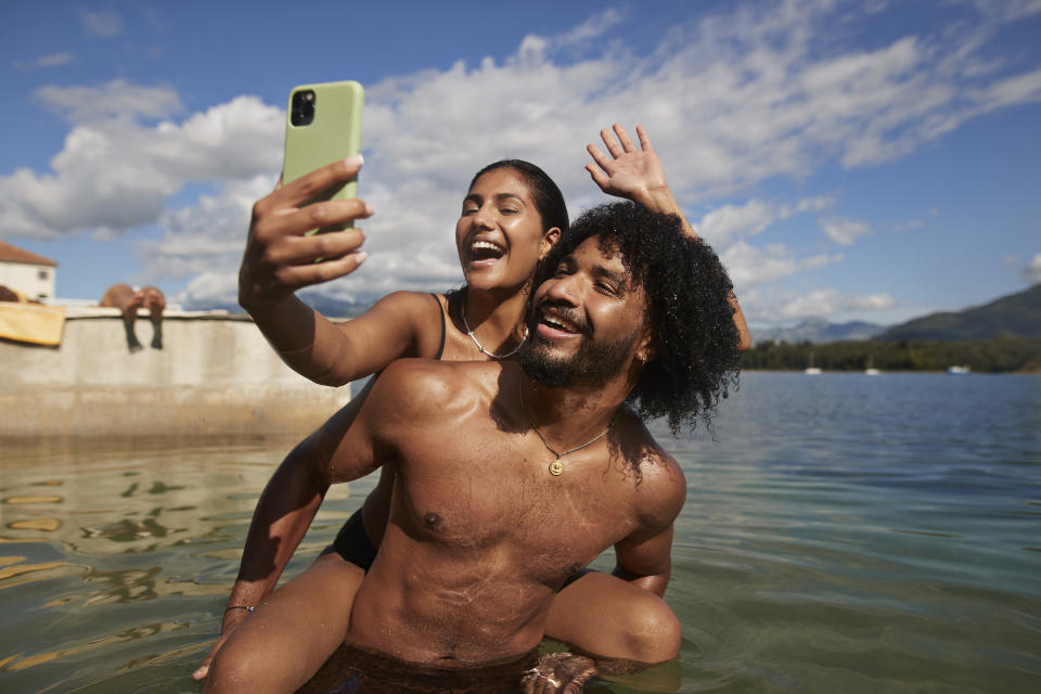 Two people take a selfie in a body of water, one standing and the other half-submerged, both smiling