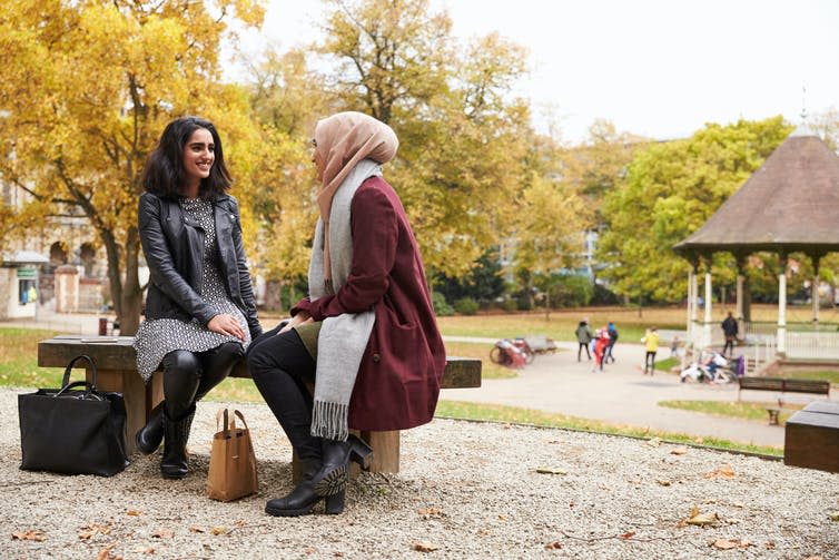 A woman in a leather jacket sits on a bench in a park with another woman in a pink hijab and marroon coat.