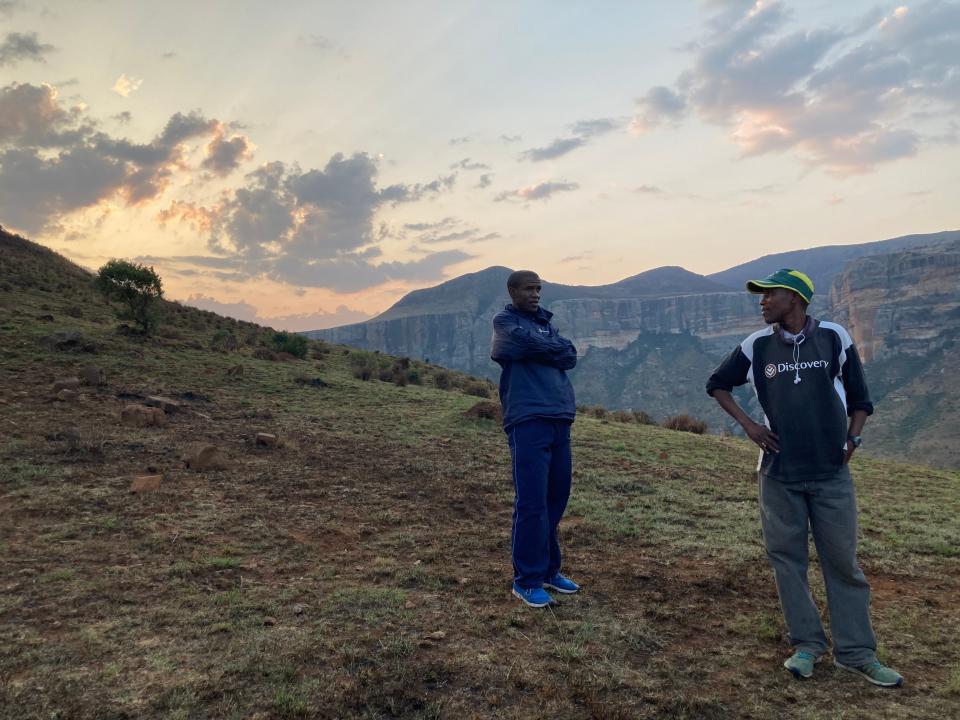 Two men stand on an incline with mountains in the background.