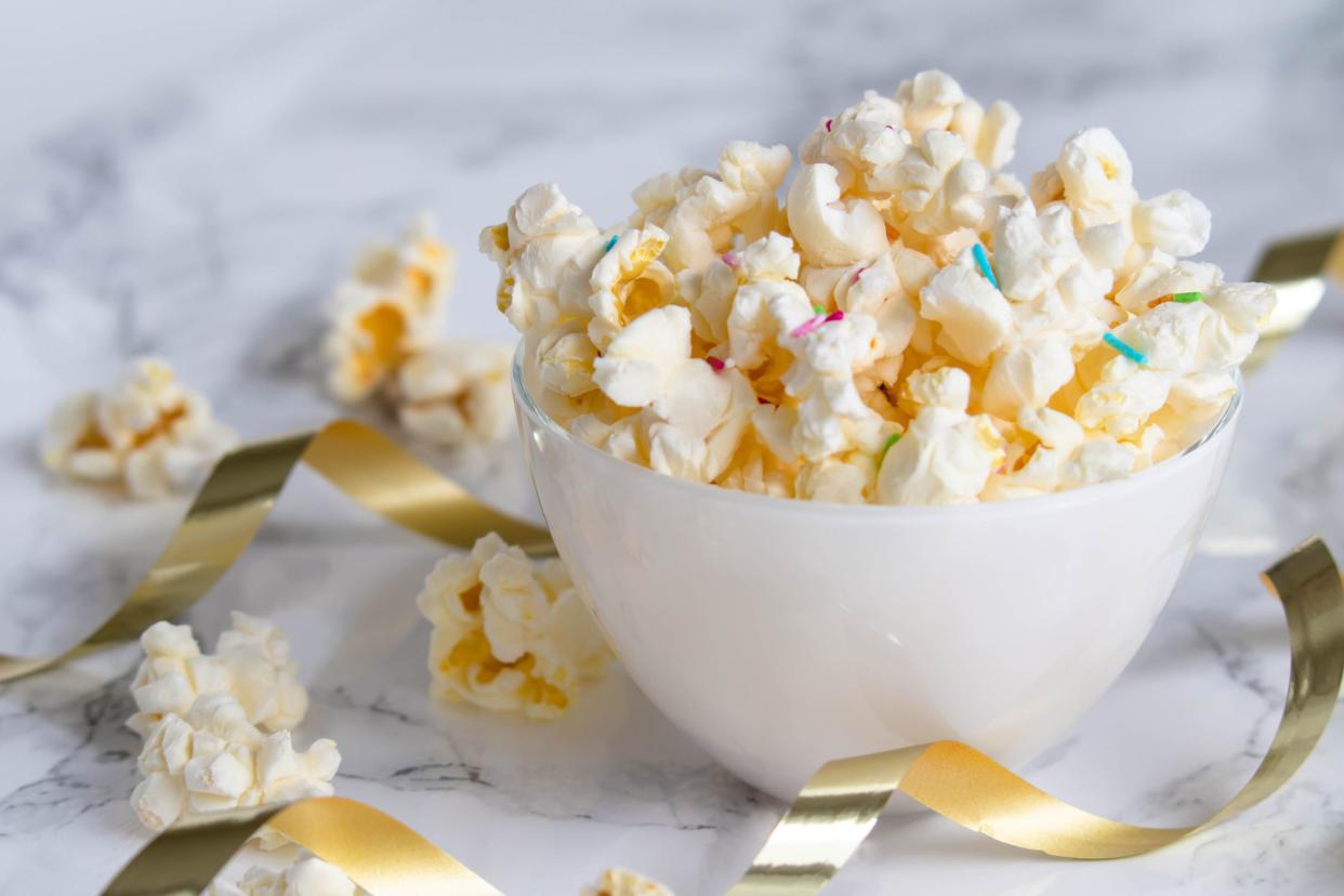 A white bowl with popcorn and colored sprinkles, on a white marble table. A few popcorns lying on the table, surrounded by golden gift ribbons.