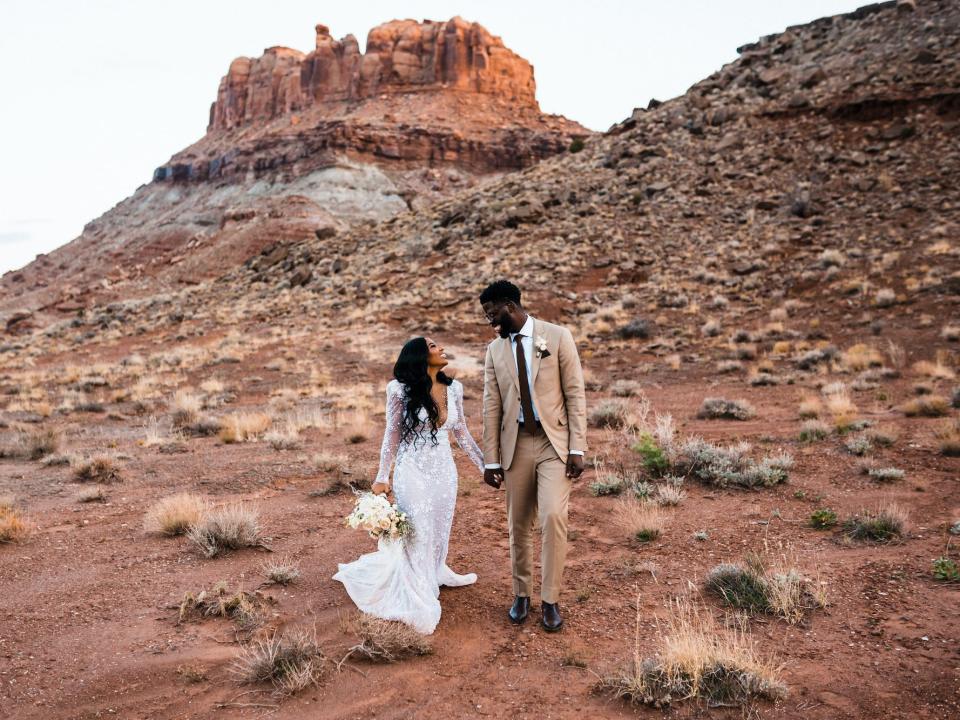 A bride and groom smile at each other in a desert in their wedding attire.