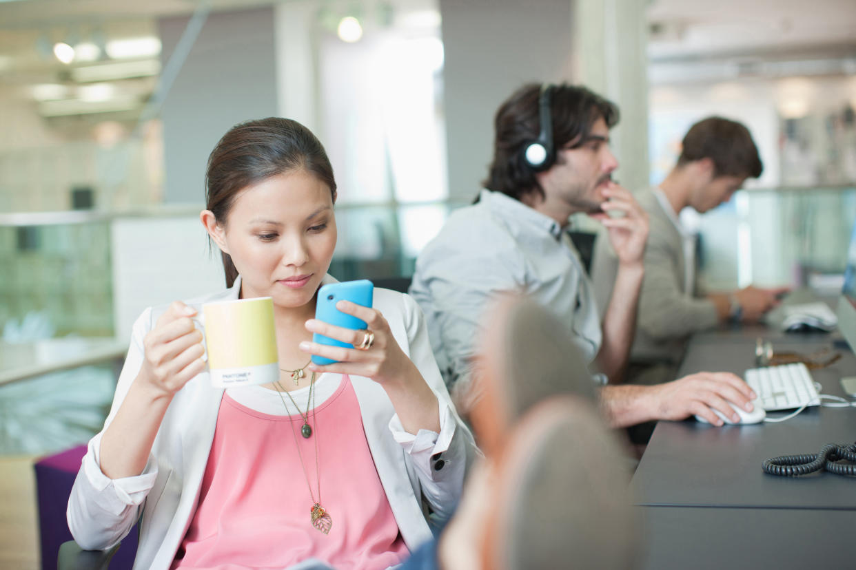 Frau mit Kaffeetasse und Smartphone, im Hintergrund arbeitende Kollegen. 