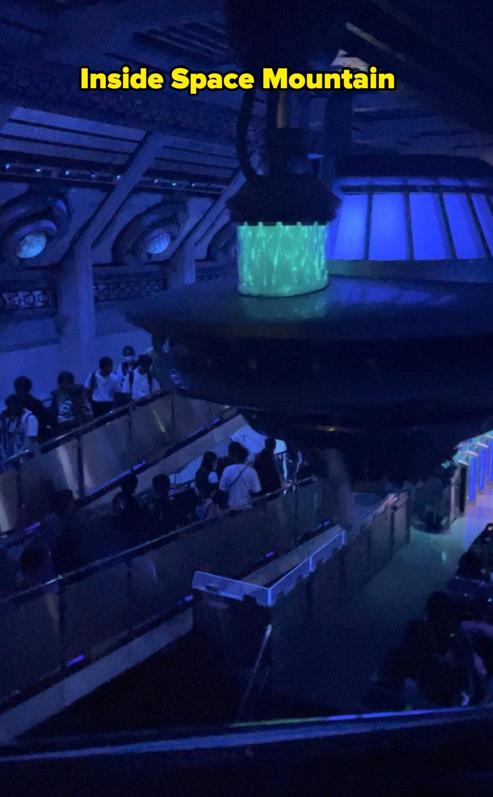 A group of people are inside an indoor theme park attraction, walking along a queue path with sci-fi themed decor and dim lighting