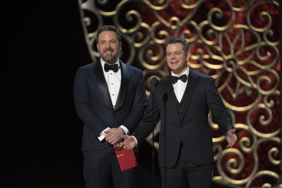 a bearded ben affleck, wearing a black tuxedo, holds a red envelope and stands next to matt damon, also wearing a black tuxedo, on a stage in front of a microphone