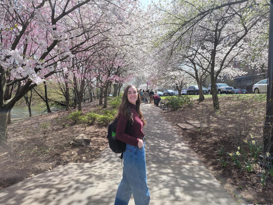 Amanda walks down a path and turns to smile at the camera. Dozens of cherry trees with blossoms ranging from white to pink line both sides of the park path.