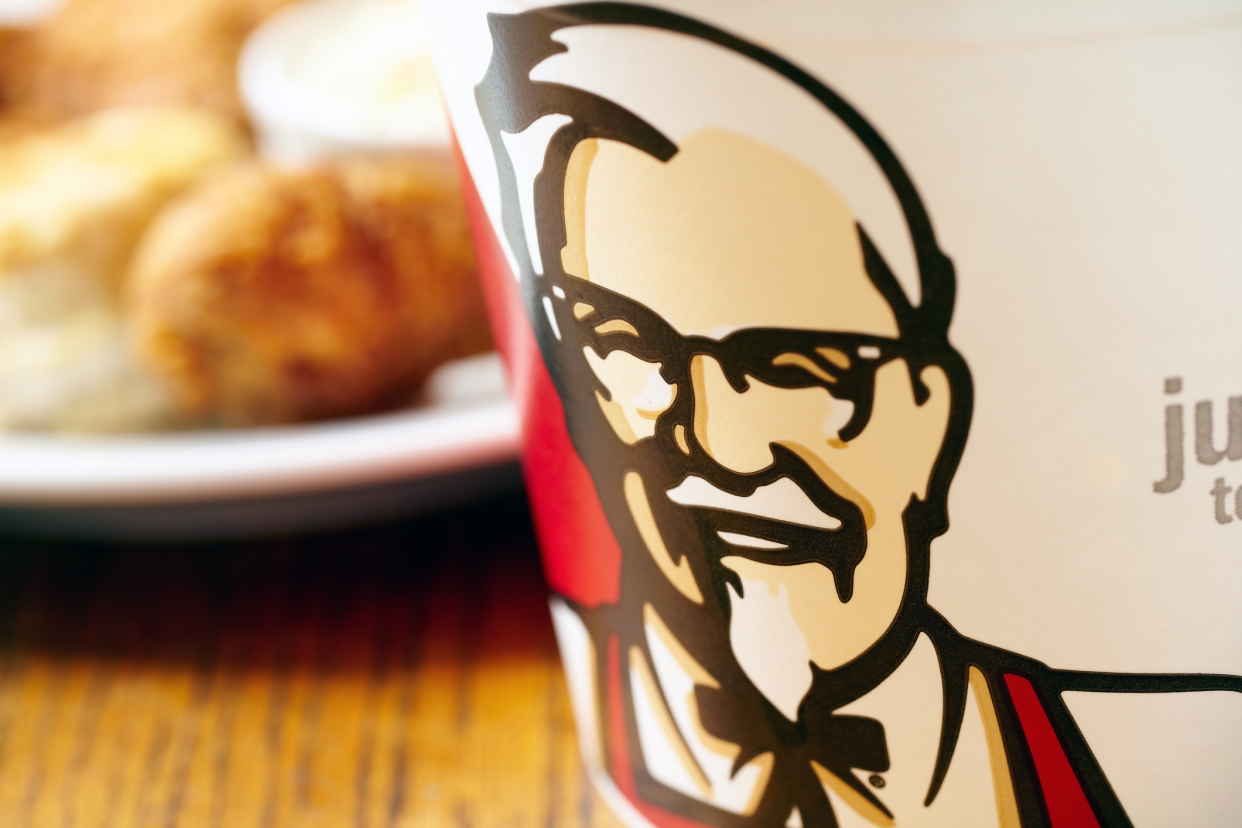 Closeup of a Kentucky Fried Chicken bucket on the right, chicken, biscuits, and mashed potatoes on a white plate blurred in the background, on a wooden table