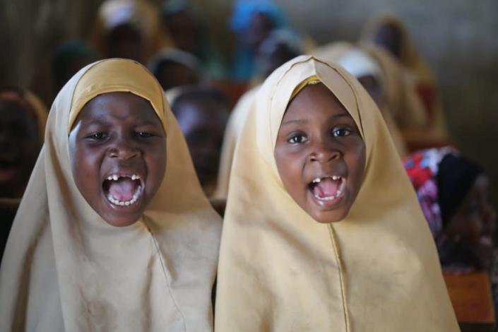 Two girls with missing milk teeth open their mouths wide as they recite in a classroom.