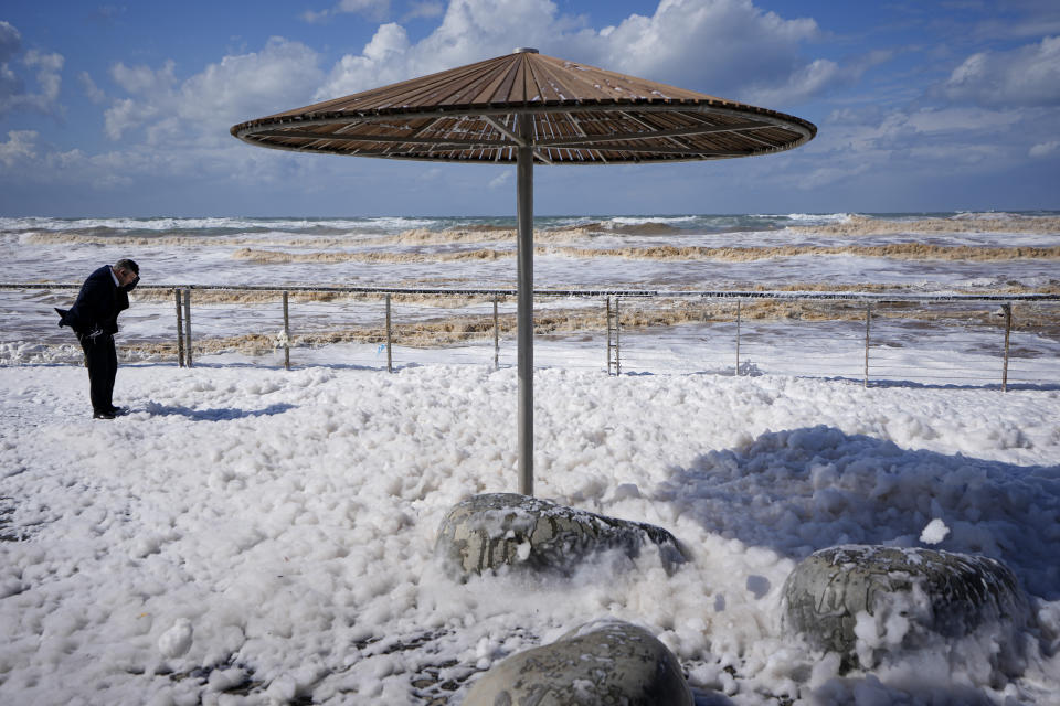 A person struggles with the wind on sea foam that covered parts of the boardwalk at the Tel Aviv's port on the Mediterranean Sea, Israel, Tuesday, Feb. 7, 2023, as strong winds and waves from Storm Barbara hit the country. (AP Photo/Ariel Schalit)