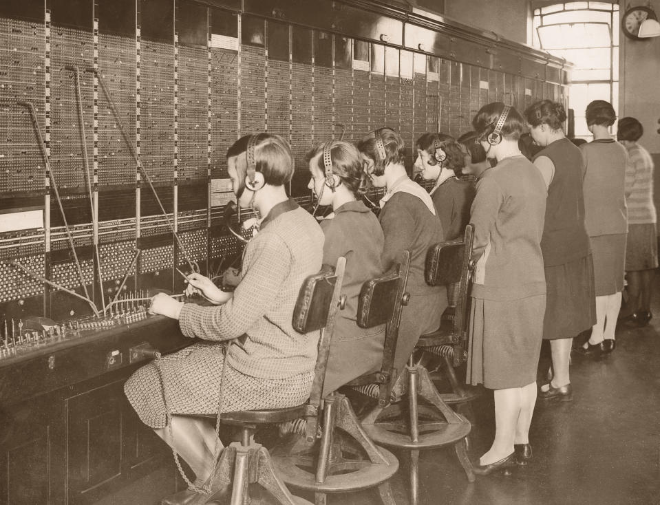 Switchboard operators sitting in chairs and manually connecting calls using a patch panel. The image depicts early 20th-century telephone communication