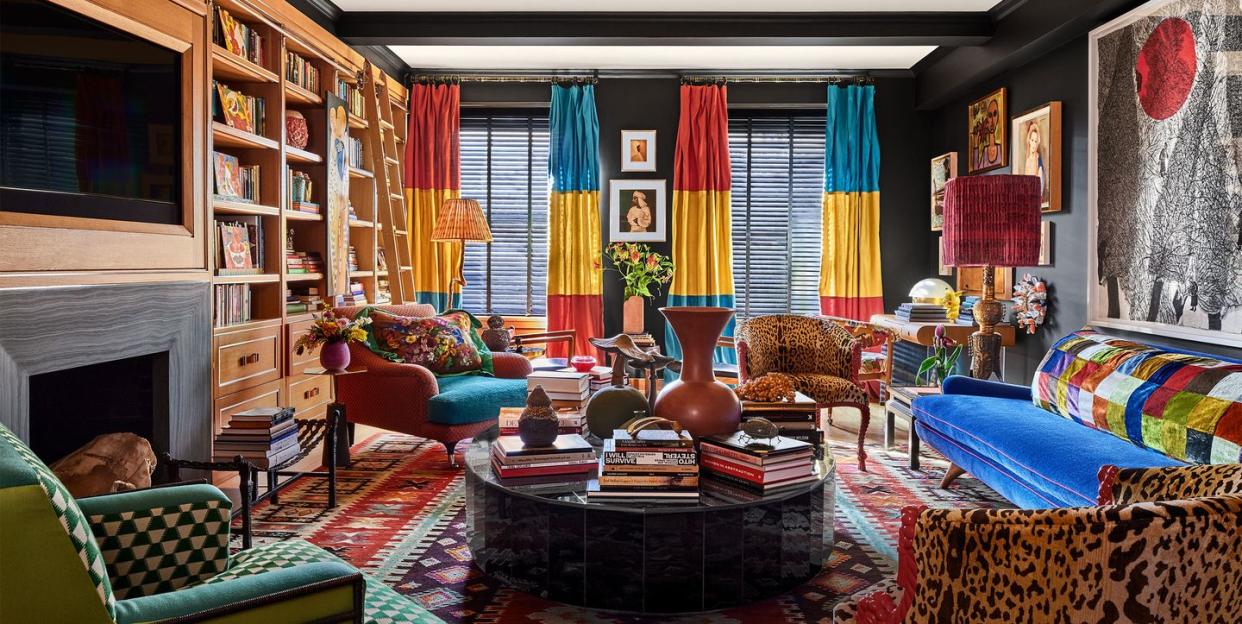 a living room has dark walls, turkish rug, bright blue sofa, club chairs with animal or geometric prints, oval cocktail tables, side tables, curtains in fuchsia, yellow, and teal, built in shelves and fireplace, large artwork above sofa