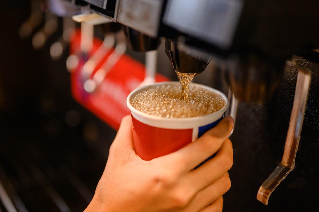 Man's hand holding a paper cup pouring a soft drink through a machine, selective focus, surrounded by a blurred background of rest of soft drink machine