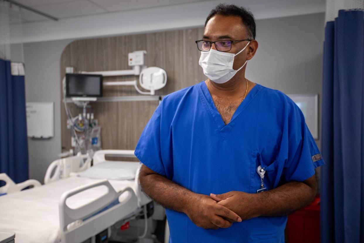 In a horizontal frame, Prasad Garimella stands in a hospital-like room, wearing scrubs and a face mask