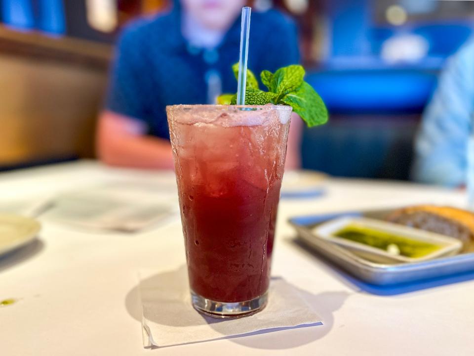 A dark-purple mocktail with mint leaves sticking out of it on a white tablecloth and napkin with a person wearing a blue shirt in the background