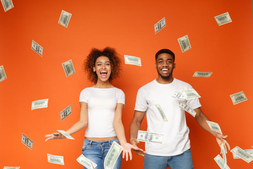 Excited young couple standing under money banknotes shower, orange background.