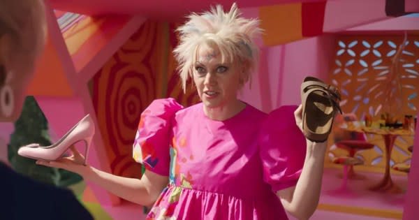 A queer Halloween costume for 2023. The image shows Kate McKinnon as Wierd Barbie. The photo is from the waist up, with a woman wearing a pink dress and short, blonde messy hair. She is holding a pink high heel in her right hand and a brown Birkenstock shoe in her left.