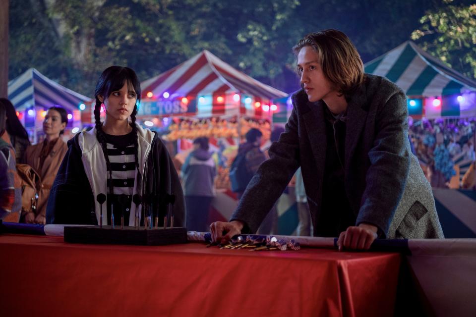 Jenna Ortega and Percy Hynes White at a carnival, standing at a game booth. Jenna wears a black-and-white striped outfit under a black cape, and Percy wears a gray coat