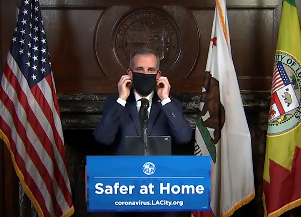 This still image taken from a live stream provided by Office of Mayor Eric Garcetti showing Los Angeles Mayor Garcetti displaying putting on a protective face mask during his daily news conference in Los Angeles on Wednesday, April 1, 2020. Garcetti has recommended that the city's 4 million people wear masks when going outside amid the spreading coronavirus. (Office of Mayor Eric Garcetti via AP)