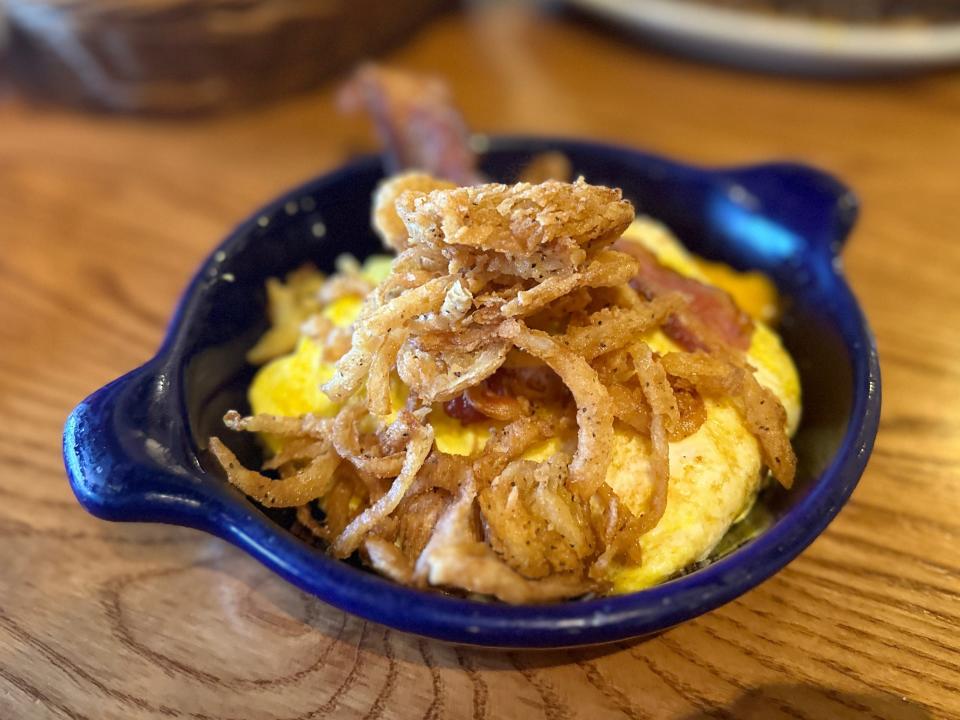 A blue bowl of scrambled eggs and pieces of crispy onions and bacon on top. The bowl sits on a wooden table.